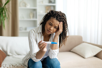 Black woman sitting on couch, worried and holding pregnancy test. African american female capturing...