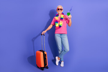 Senior woman in casual striped outfit holding a drink and suitcase ready for vacation against...