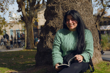 Portrait of happy young woman with a book sitting in the park looking at the camera