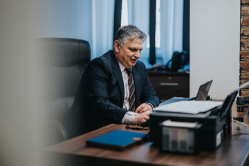 A professional businessman in a suit sits smiling behind a desk in a modern office. Documents and...