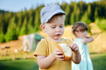 Young boy in yellow shirt and blue cap intently examines bun in hands while standing in grassy...