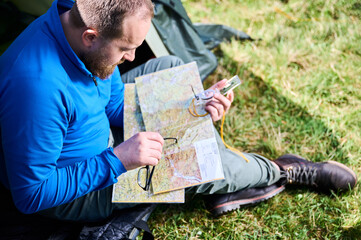Close up man in glasses closely examines map while holding compass. He sits by his tent, deeply...