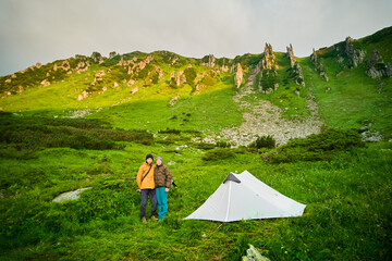 Two campers stand next to white tent, surrounded by lush green hills and rocky cliffs. Dramatic...