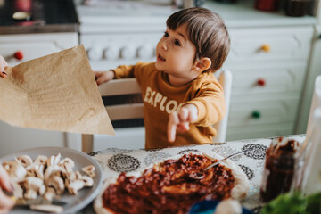 A young boy wearing a colorful sweater assists in making a homemade pizza by spreading sauce and...