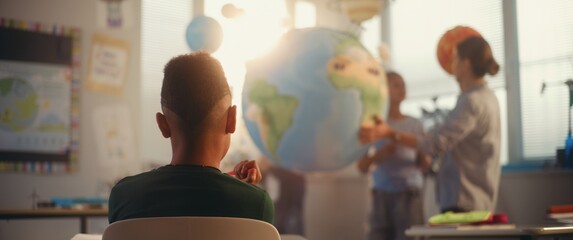 STEM Class on Renewable Energy: Female Teacher Explaining Energy Resources to Elementary School Students Using Earth Model. Rack Focus on African American Boy Learning Science in Classroom. Dolly Shot