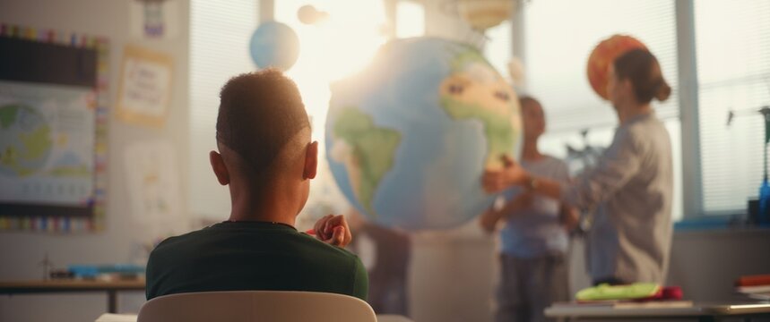 STEM Class on Renewable Energy: Female Teacher Explaining Energy Resources to Elementary School Students Using Earth Model. Rack Focus on African American Boy Learning Science in Classroom. Dolly Shot - Powered by Adobe