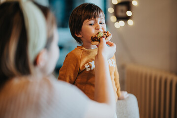 A toddler is being fed by their mother while sitting in a cozy, indoor home environment. Warm...