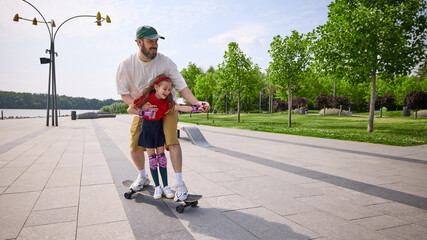Speed and laughter. Father instructs his daughter in skateboarding along a sunlit park pathway,...