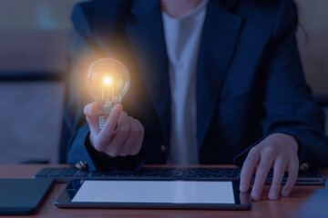 A businesswoman is using a keyboard to work on a wooden table while holding a light bulb. Have an...