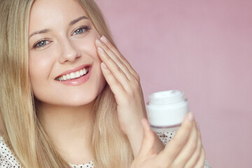 Smiling blonde woman applying moisturiser, holding face cream jar. Clear skin, blue eyes, beauty...