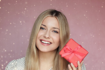 Smiling young woman with blonde hair and blue eyes holding a red gift box with a ribbon, pink...