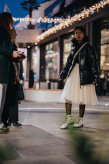 Stylish young woman dressed in white and black ensemble standing on an urban street. The nighttime...