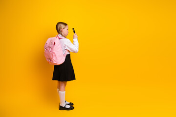 Young schoolgirl with backpack exploring with a magnifying glass on yellow background, embracing education and curiosity