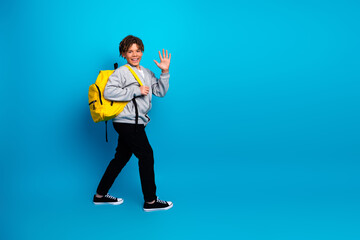 Smiling young boy with yellow backpack waves against a bright blue background, showcasing school...