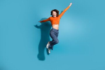 Cheerful young female in orange outfit jumping joyfully on a vibrant blue background expressing...