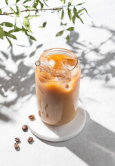 Ice latte coffee in a glass with milk on a light background with coffee beans, branch and morning shadows.