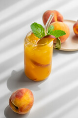 Ice tea with peach, mint in a glass on a white table near window with fresh fruits and shadow.