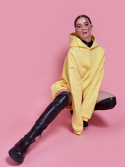 A woman in a yellow hoodie and black boots poses for a photograph. The photo is bright and...