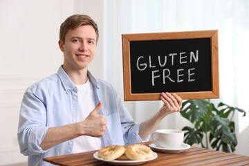 Man holding small chalkboard with text Gluten Free and showing thumbs up at wooden table indoors
