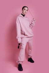 A woman in a pink hoodie and sweatpants poses for a photo. The pink color of her outfit gives off a...