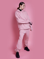 A woman in a pink hoodie and sweatpants poses for a photo. The pink color of her outfit gives off a...