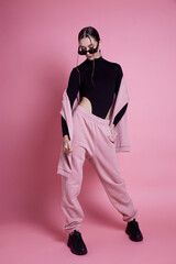 A woman in a pink hoodie and sweatpants poses for a photo. The pink color of her outfit gives off a...