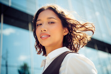 Beautiful young businesswoman standing outdoors by a modern glass building with charming expression...
