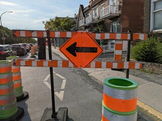 Bright orange construction sign with a left-pointing black arrow stands amid traffic barrels,...