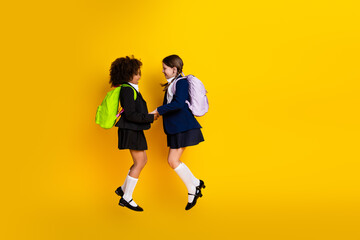 Two cheerful schoolgirls smiling and jumping together on a vibrant yellow background wearing school uniforms and backpacks