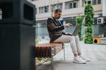 A businessman sits on a wooden bench in an urban setting, utilizing his laptop while gesturing towards the screen, with green plants and modern architecture around.