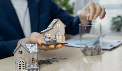 Businessman placing coin into jar while holding a small house model, symbolizing real estate...