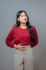 An Indonesian woman in a red shirt and beige pants, looking up while holding her stomach, showing a...
