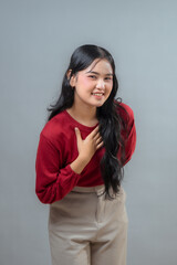An Indonesian woman in a red shirt and beige pants, smiling while placing her hand on her chest....