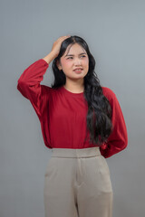 A thoughtful Indonesian woman in a red top and beige pants scratching her head with a confused...