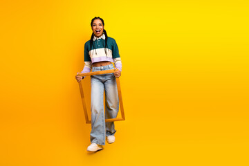 Fototapeta premium Smiling young woman in casual outfit holding wooden frame on vibrant yellow background, showcasing stylish and trendy look