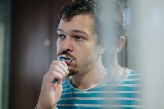 A young man dressed in a striped shirt brushing his teeth while looking into a mirror, emphasizing dental care and personal hygiene routines.