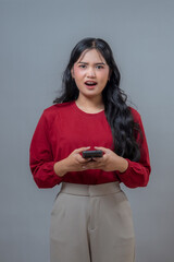 An Indonesian woman in a red blouse holding a smartphone looks shocked and surprised, reacting...