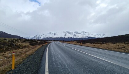 Fototapeta premium Scenic road stretches toward snow-capped mountains under a cloudy sky, inviting exploration and offering a sense of adventure in the landscape.