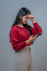 An Indonesian woman in a red top and beige pants looks frustrated and stressed while holding a...