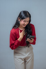 An Indonesian woman in a red blouse and beige pants smiles warmly while looking at her smartphone,...