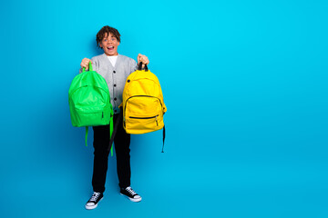 Energetic boy holding two colorful backpacks standing against a vibrant blue background, smiling...