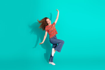 Young stylish woman jumps joyfully on a teal turquoise background, wearing a striped t-shirt,...