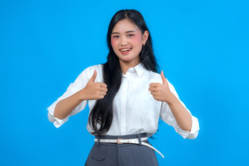 Fototapeta premium Portrait of a cheerful young Asian woman showing two thumbs up with a confident smile. Dressed in business casual, she stands against a solid blue background conveying positivity and approval.