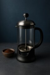 French press coffee maker with coffee grounds in a small bowl beside it. Dark background enhances the coffee tones.