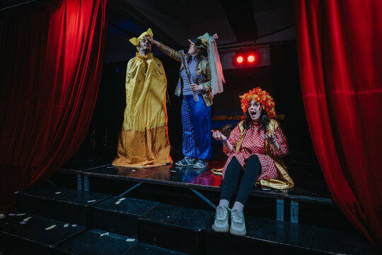 Two actors dressed in colorful costumes perform an engaging theatrical comedy scene on a decorated stage, capturing lively expressions and dramatic ambience under stage lights.
