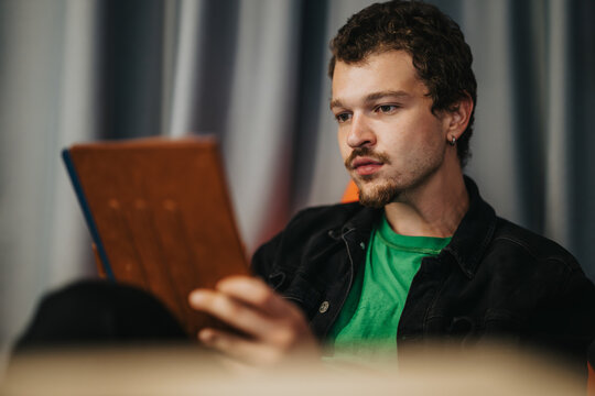 Teen boy dressed casually in a green shirt and denim jacket, sitting indoors against a backdrop, and reading a tablet while concentrating. The scene conveys a sense of focus and quiet engagement. - Powered by Adobe