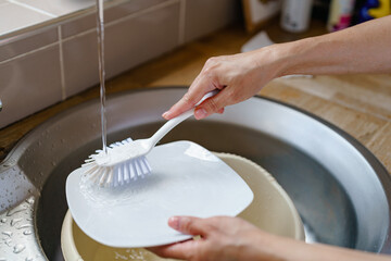 Washing dishes at a kitchen sink while water flows from a faucet and hands hold a dish sponge and a plate in an evening setting