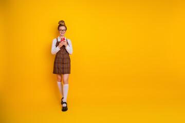Young female student in fashionable attire holding smartphone on vibrant yellow background, conveying vibrant youthfulness