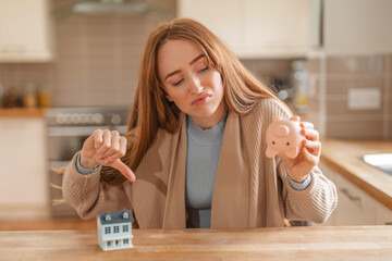 Woman shows disappointment while comparing small house model with piggy bank in a modern kitchen...