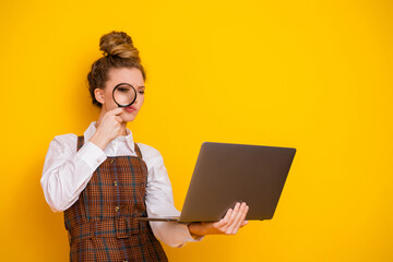 Young woman in stylish attire inspecting laptop closely with magnifying glass against bright yellow...